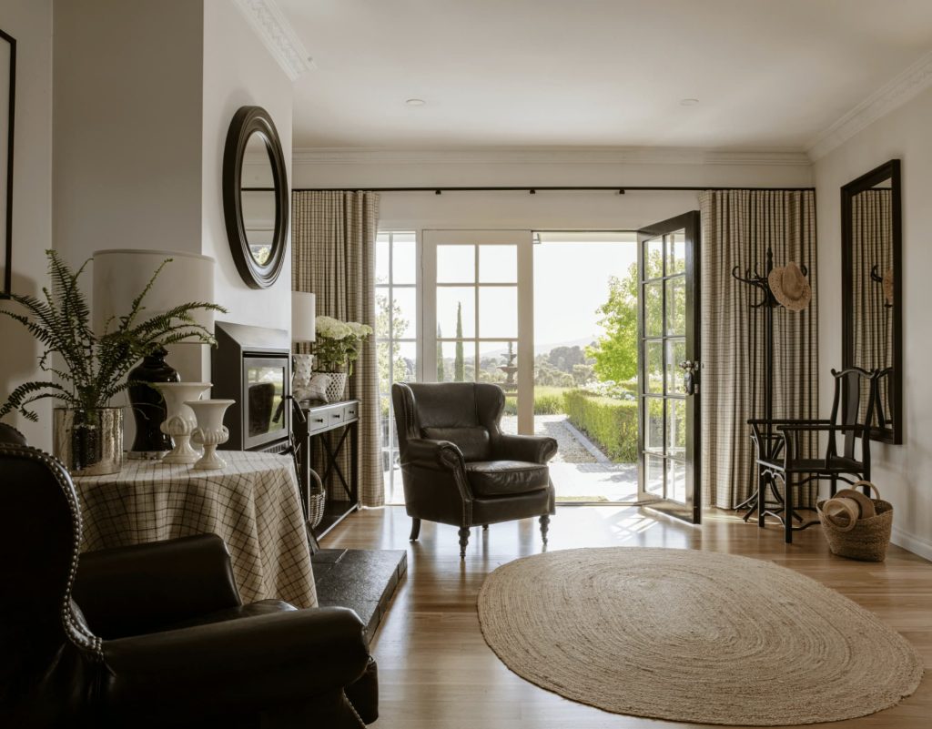 A cozy living room with leather chairs, a round rug, and a table. A door opens to a garden view. Walls are decorated with mirrors and a plant is on a table. Light filters through curtains.