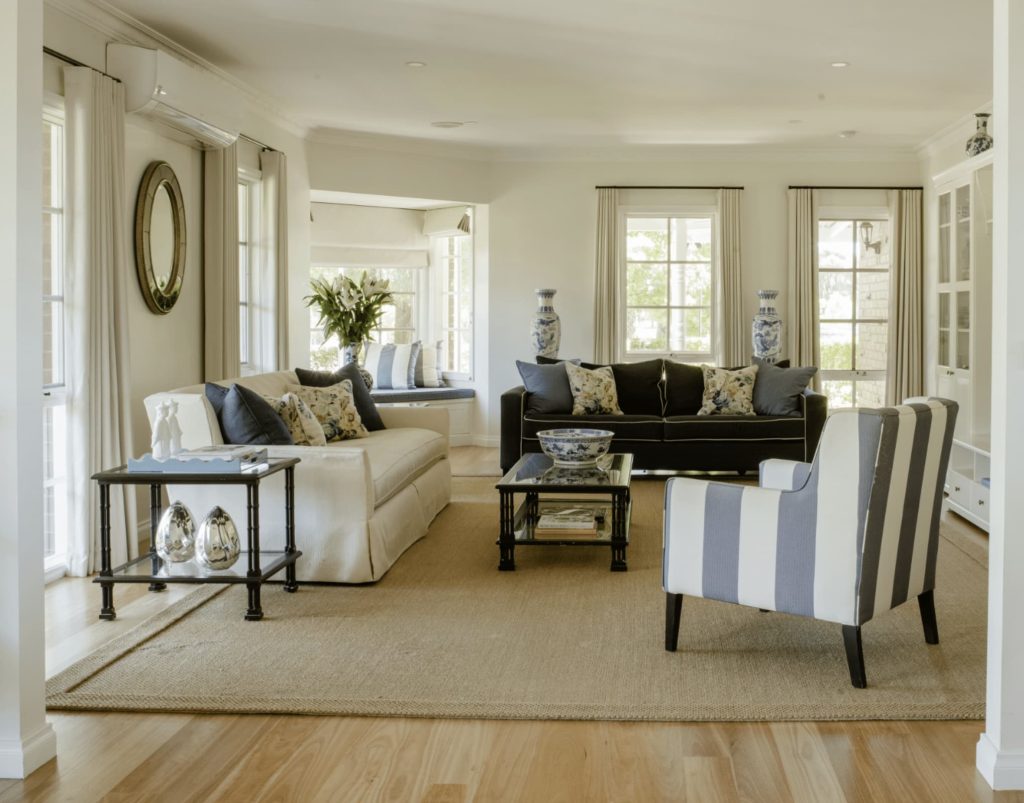 A modern living room with white and black sofas, a striped armchair, glass table, decorative vases, and a beige rug on wooden floors. Large windows provide natural light.