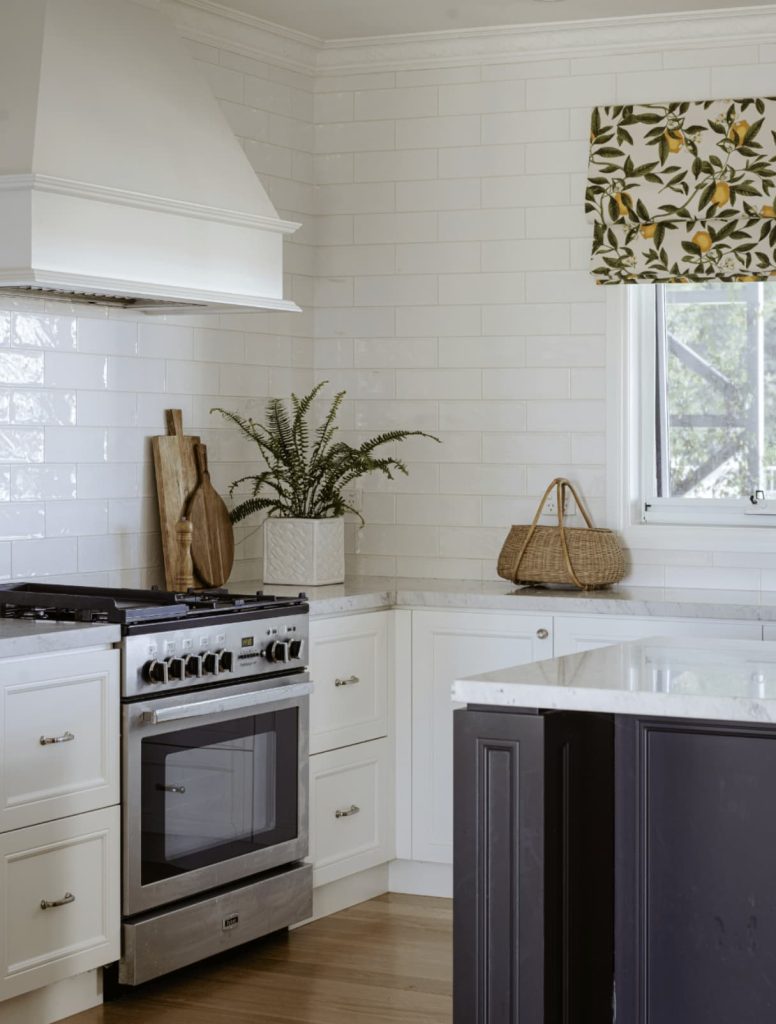 A white kitchen with a stove, wooden cutting boards, a potted plant, and a wicker basket. The counters are marble, and there's a window with a floral curtain.