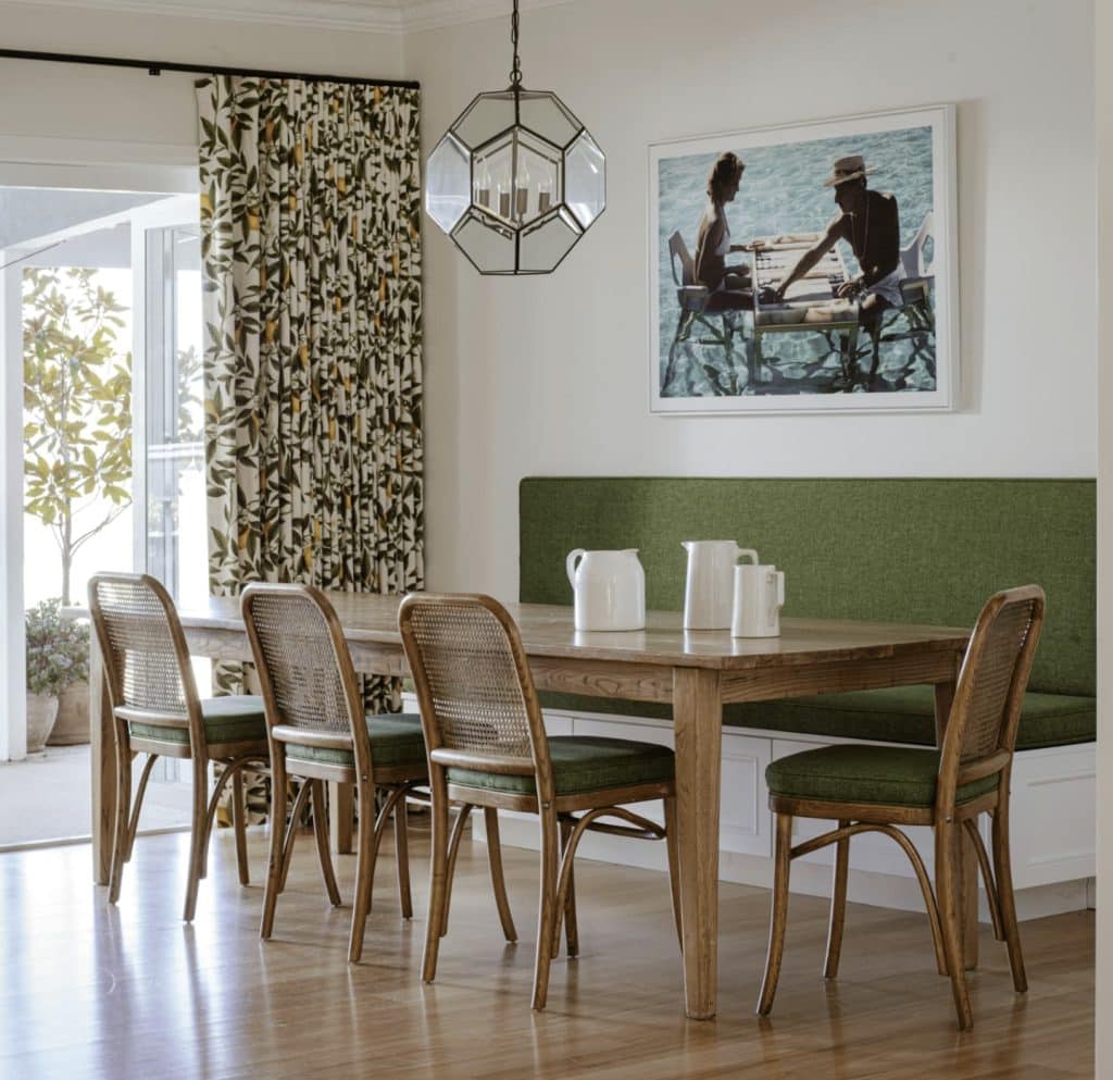 Dining room with a wooden table, green cushioned chairs, and a bench. A geometric light fixture hangs above, with patterned curtains and a framed picture on the wall. Sunlight streams in.