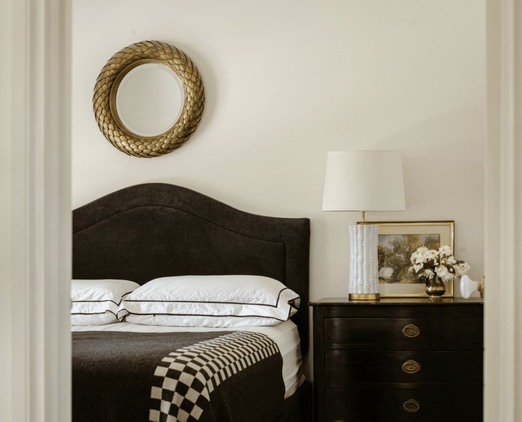 Cozy bedroom with a dark bed, white pillows, and a checkered blanket. A round mirror hangs above the bed. A lamp and framed picture sit on a dark bedside table with flowers.