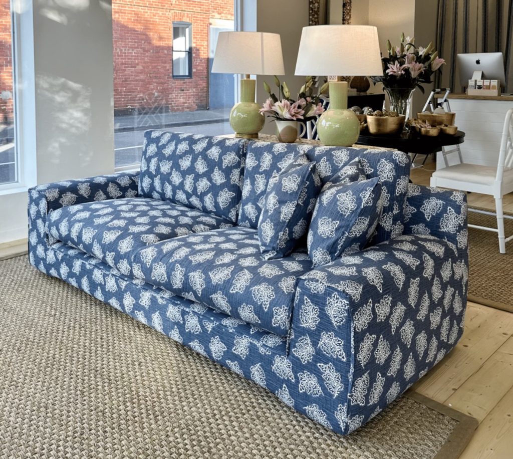 A blue and white patterned sofa with matching pillows is placed on a woven rug in a well-lit room. Two green lamps and pink flowers are on a nearby table.