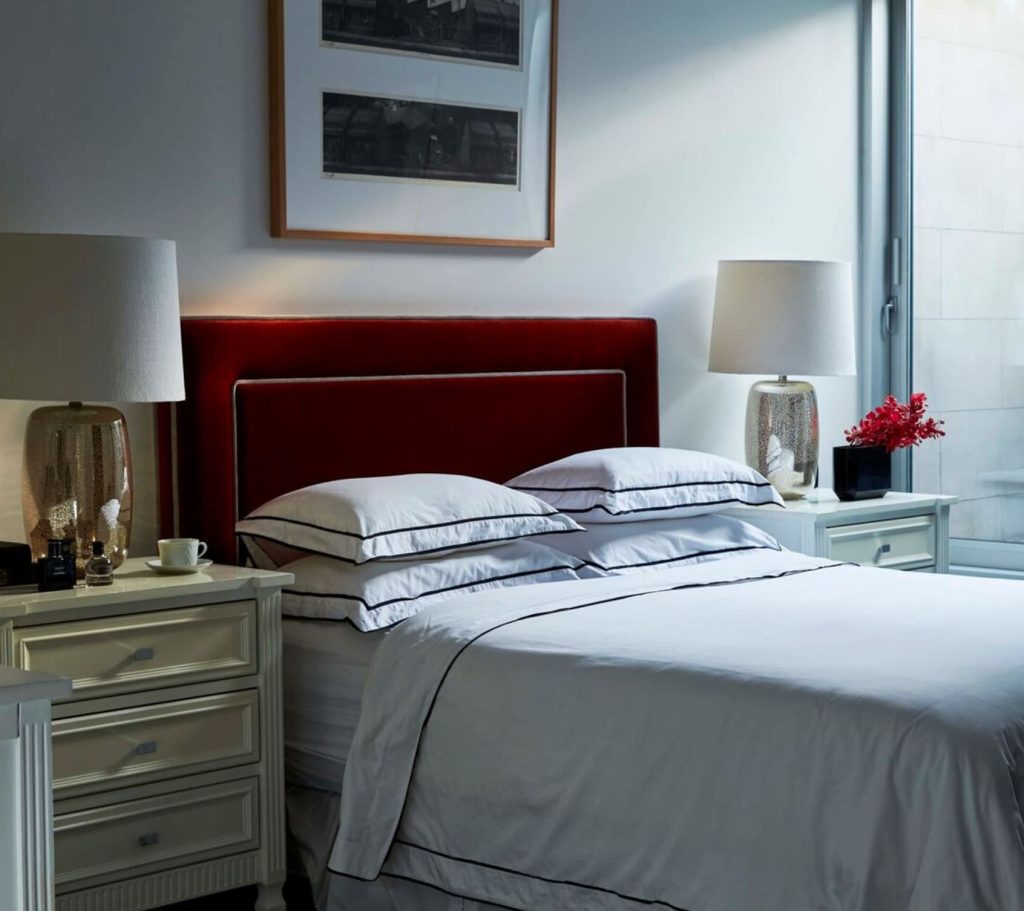 A neatly made bed with white linens and black trim, flanked by two bedside tables with lamps. Red cushioned headboard, framed artwork above, and a vase with red flowers on the right table.