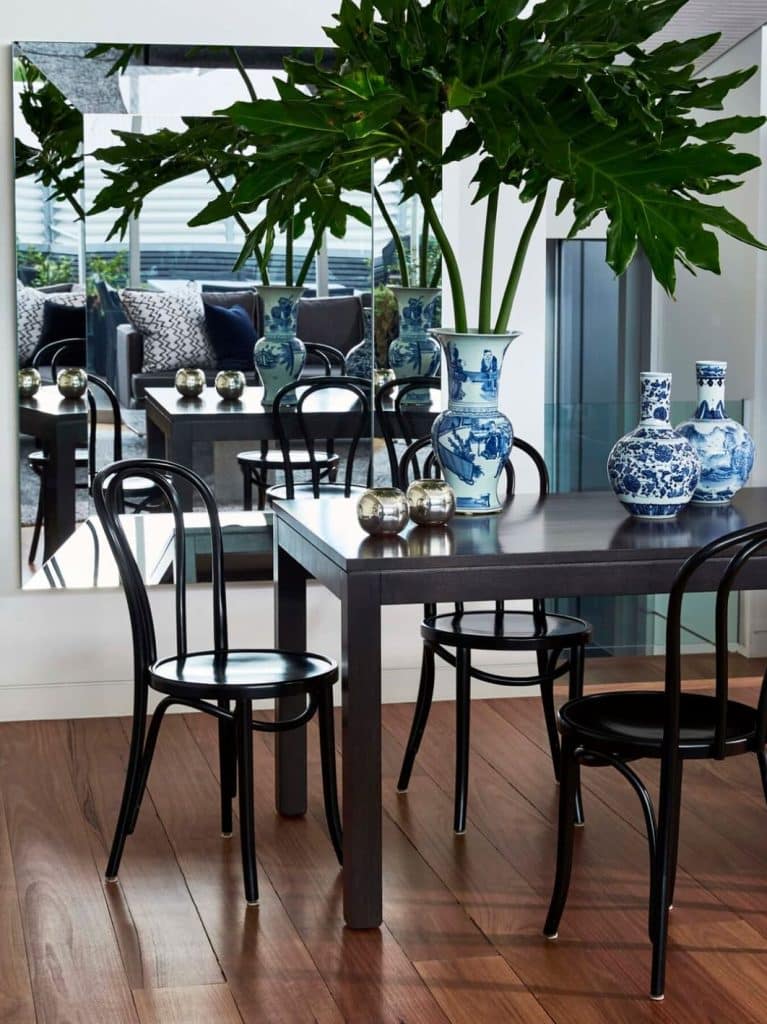 Dining area with a dark table, black chairs, blue and white vases, and plants. Large mirror reflects the scene, with outdoor seating visible.