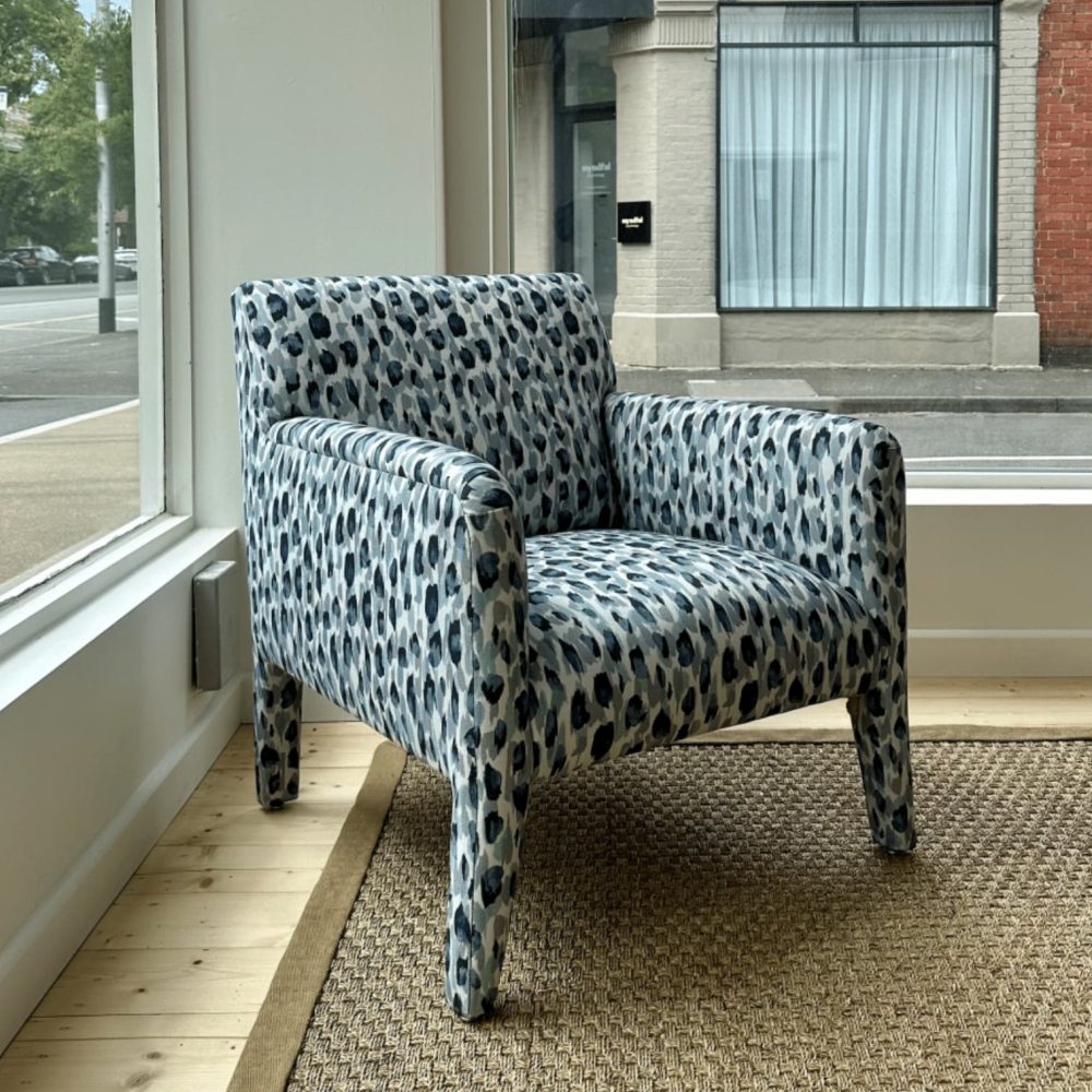 Leopard-print upholstered armchair with a boxy design placed on a woven rug in a room with large windows.